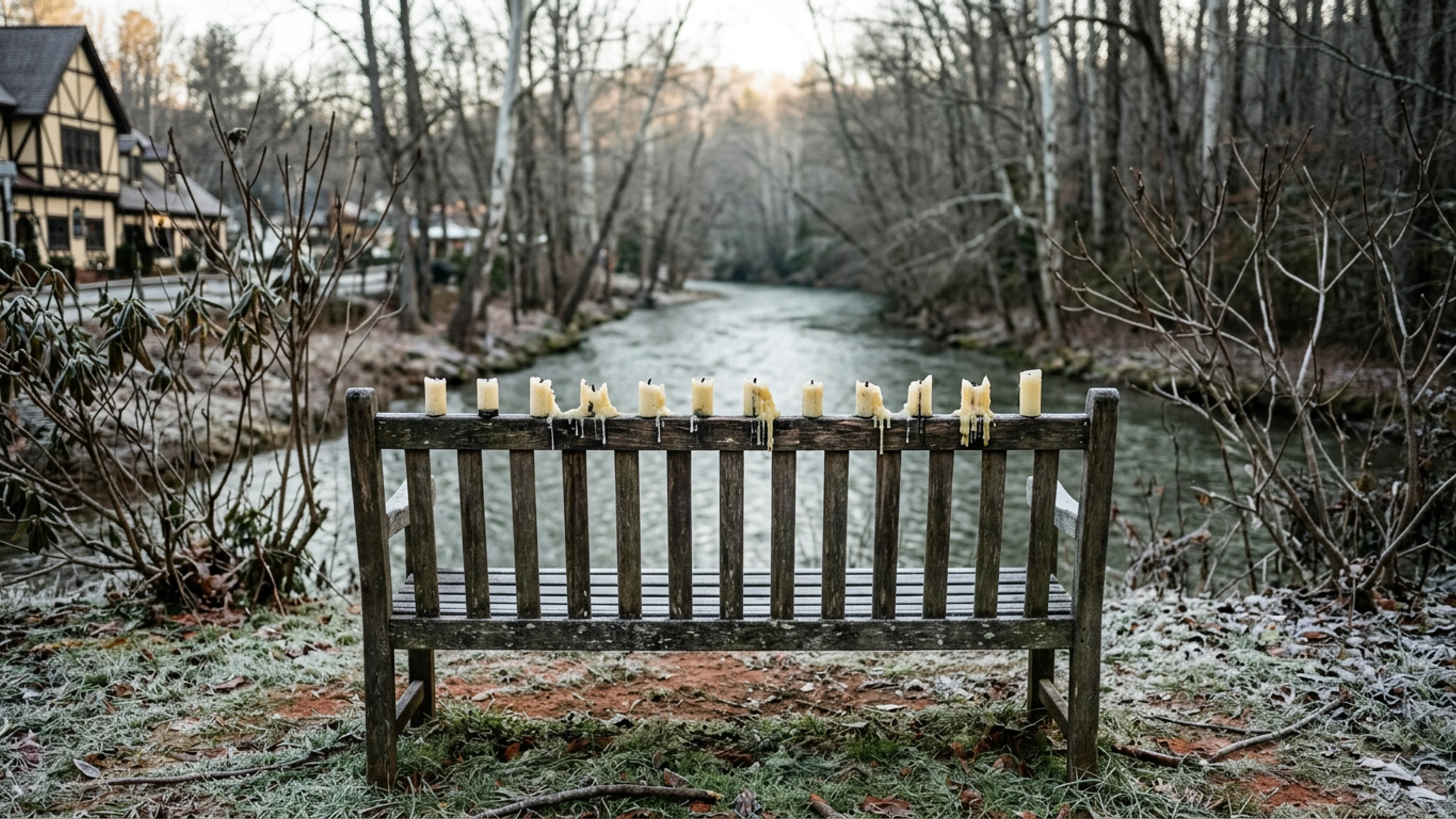 The small wooden park bench on Bruckenstrasse, between the Helen Welcome Center and the Chattahoochee pedestrian bridge, photographed Friday morning, twelve hours after the Thursday-evening vigil. Twelve candle-stubs are visible, arranged in a single row along the bench's back rest. The river, visible in the distance, is gray-green and still. (Photo: Bavarian Brainrot / Edmund Crowe)
